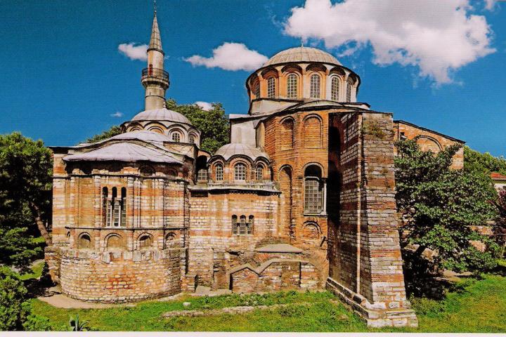 a large brick building with grass in front of a castle with Chora Church in the background
