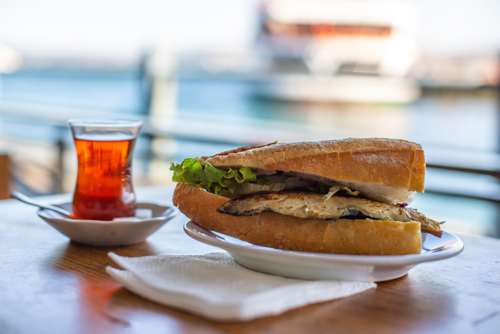 Fish sandwich and a glass of tea on a wooden table beside a waterfront view.