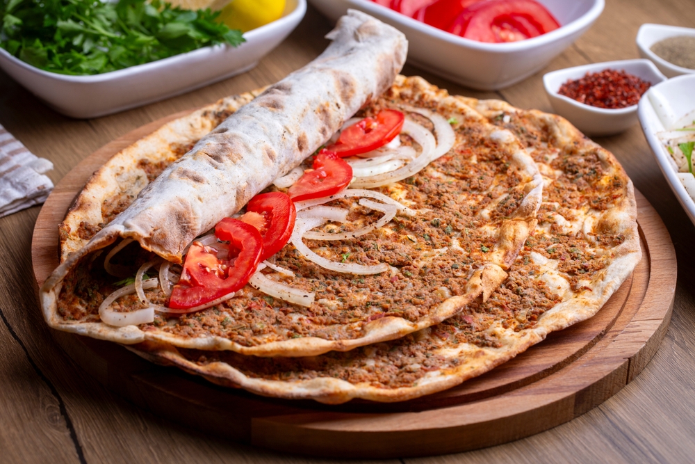Two Lahmacun with toppings of tomato and onion on a wooden board with herbs and spices in the background.