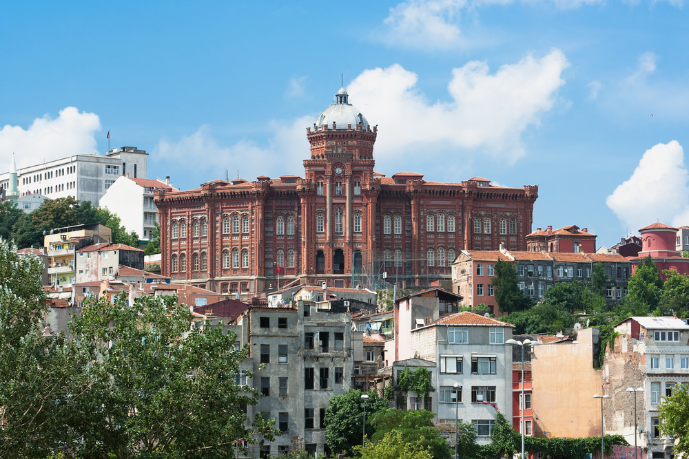 Historic red building on a hill, surrounded by smaller buildings under a blue sky with clouds.